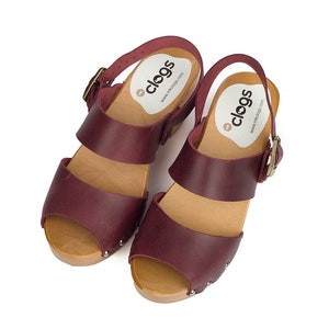 Puede incluir: Un par de sandalias de tac&oacute;n de cuero burdeos con plataforma de madera y cierre de hebilla. Las sandalias tienen un dedo del pie abierto y est&aacute;n decoradas con peque&ntilde;os tachuelas plateadas. Las sandalias tienen una etiqueta que dice "clogs" y una direcci&oacute;n de sitio web.