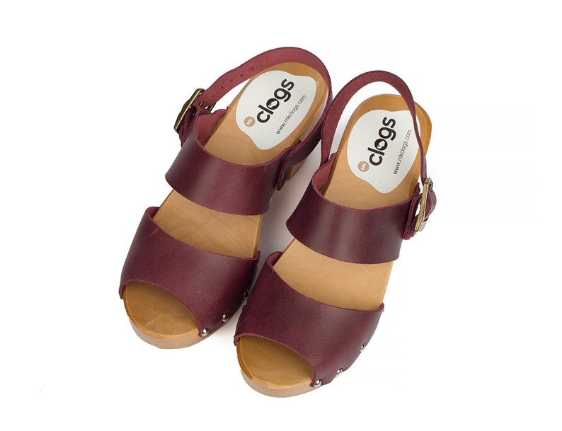 Puede incluir: Un par de sandalias de tac&oacute;n de cuero burdeos con plataforma de madera y cierre de hebilla. Las sandalias tienen un dedo del pie abierto y est&aacute;n decoradas con peque&ntilde;os tachuelas plateadas. Las sandalias tienen una etiqueta que dice "clogs" y una direcci&oacute;n de sitio web.