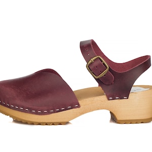May include: Burgundy leather clog sandal with a wooden sole. The sandal features a closed toe, an adjustable ankle strap with a brass buckle, and white stitching details. The heel is approximately 2 inches high.