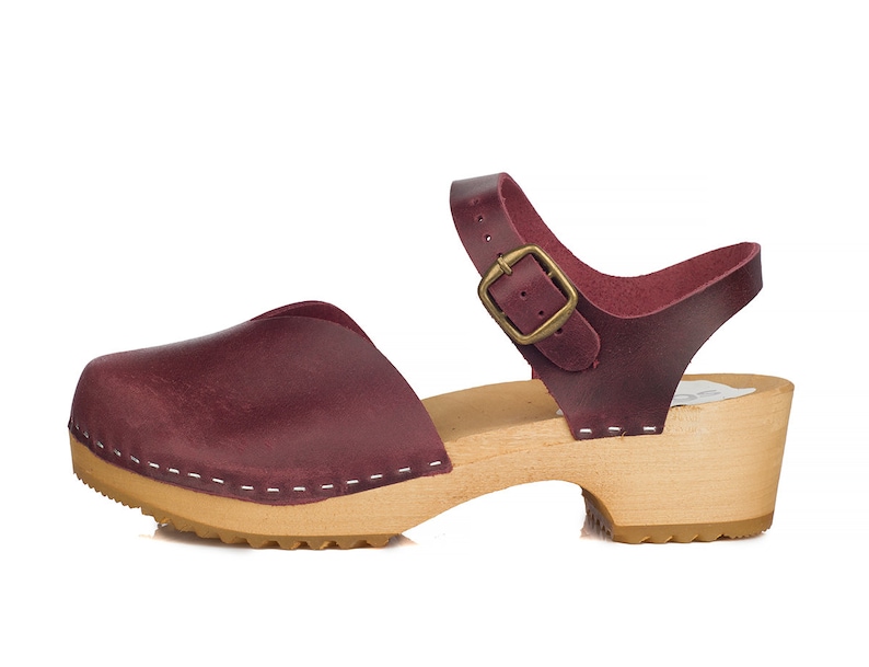 May include: Burgundy leather clog sandal with a wooden sole. The sandal features a closed toe, an adjustable ankle strap with a brass buckle, and white stitching details. The heel is approximately 2 inches high.