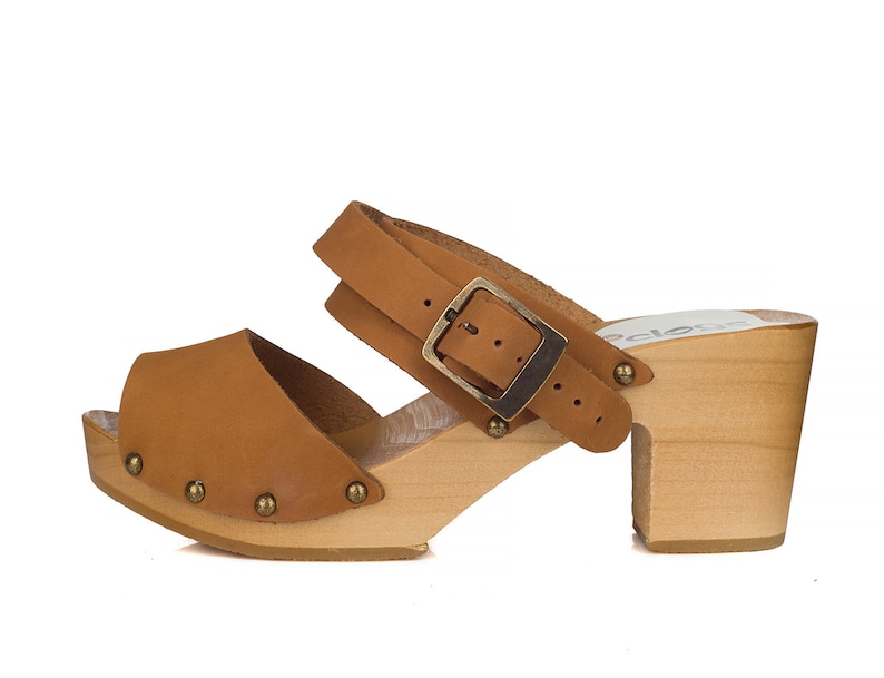 Puede incluir: Sandalias de madera con tac&oacute;n de cuero marr&oacute;n claro con una correa de hebilla y una plataforma de madera. Las sandalias tienen peque&ntilde;os tachuelas met&aacute;licas alrededor del borde de la suela.