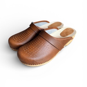 May include: A pair of brown leather clogs with a perforated design on the toe box. The clogs have a strap across the top and a light-colored sole. The shoes are set against a white background.