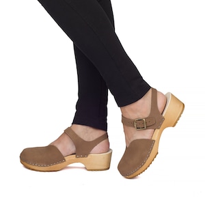 May include: Brown suede clogs with a wooden sole. The clogs have a closed toe and an ankle strap with a buckle. The sole is made of light-colored wood. The clogs are worn with black pants.