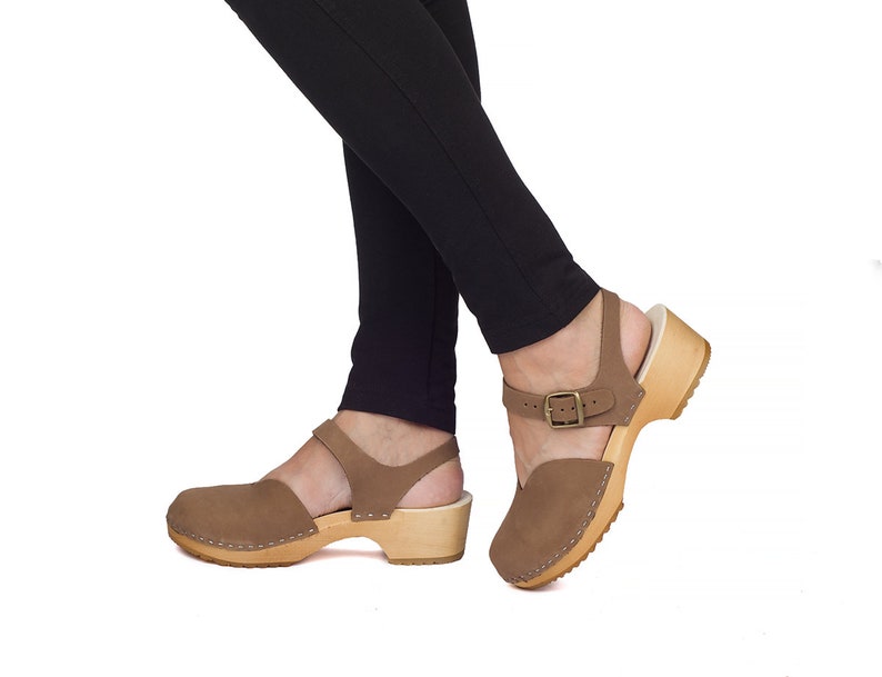 May include: Brown suede clogs with a wooden sole. The clogs have a closed toe and an ankle strap with a buckle. The sole is made of light-colored wood. The clogs are worn with black pants.