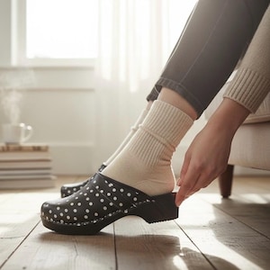 May include: A black clog with a white polka dot pattern, featuring a rounded toe, a sturdy sole, and a strap across the top. The shoe is worn with a beige ribbed knit sock, providing a contrast to the dark, patterned exterior of the footwear.