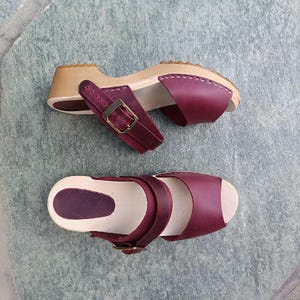 May include: A pair of burgundy leather clogs with a wooden base and buckle closures. The clogs have a peep-toe design and are made of a soft, supple leather.