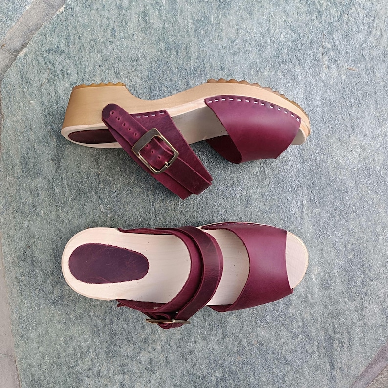 May include: A pair of burgundy leather clogs with a wooden base and buckle closures. The clogs have a peep-toe design and are made of a soft, supple leather.