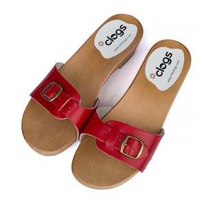 May include: A pair of red leather clogs with a buckle closure. The clogs have a wooden base and are made by the company "clogs".