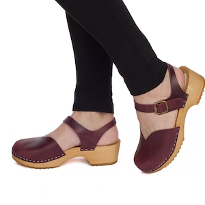 May include: Burgundy leather clogs with a wooden sole and ankle strap. The clogs have a closed toe design and a buckle closure. The wooden sole is light brown and the leather is a deep burgundy color. The clogs are shown on a white background.