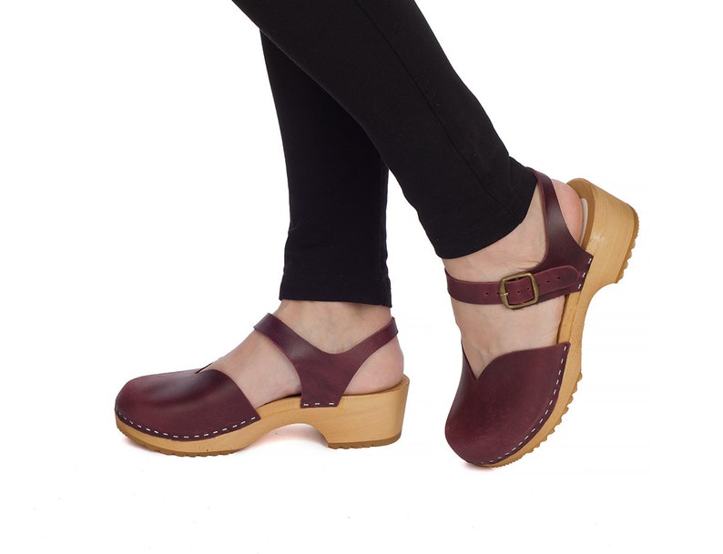 May include: Burgundy leather clogs with a wooden sole and ankle strap. The clogs have a closed toe design and a buckle closure. The wooden sole is light brown and the leather is a deep burgundy color. The clogs are shown on a white background.
