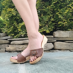 May include: Brown leather clog sandals with a buckle fastening and a wooden platform sole. The sandals have a dark brown leather strap that goes over the foot and a buckle fastening on the top of the foot. The sandals have a wooden platform sole that is about 5 cm high.