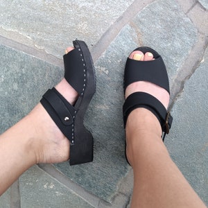 May include: A pair of black leather sandals with adjustable straps and a closed toe. The sandals have a wooden sole and are worn on a person's feet.