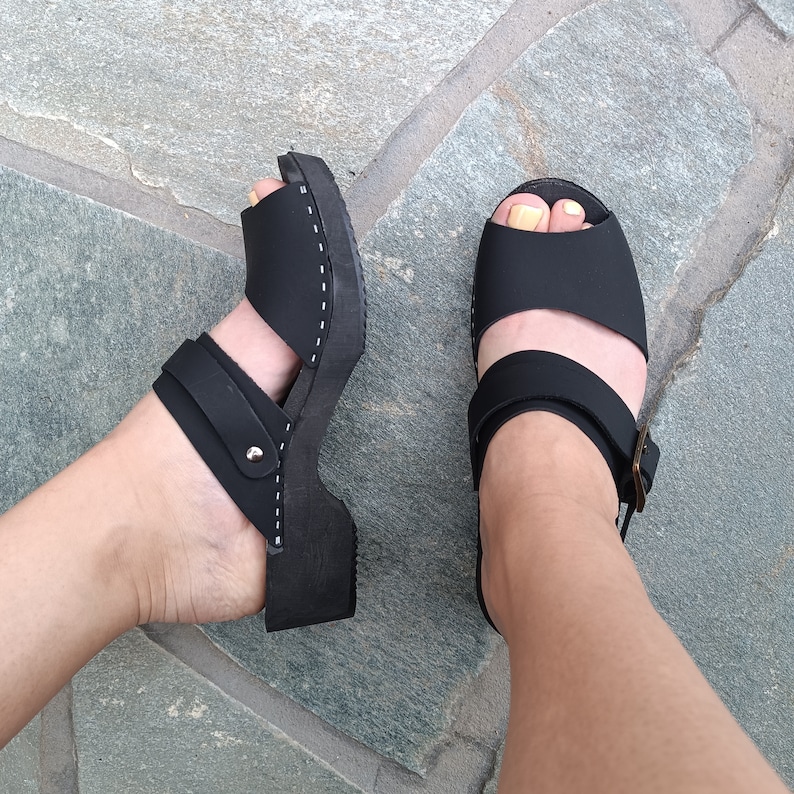May include: A pair of black leather sandals with adjustable straps and a closed toe. The sandals have a wooden sole and are worn on a person's feet.