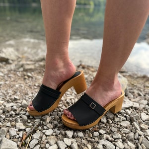 May include: A pair of black leather clogs with wooden heels. The clogs have a buckle on the strap and are being worn by someone standing on a gravel path.