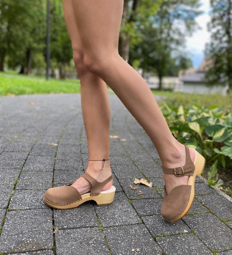 May include: Brown suede clogs with a wooden sole and a strap across the top of the foot and around the ankle. The clogs have a buckle closure and are worn outdoors on a paved surface.