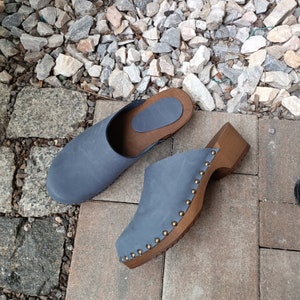 Könnte beinhalten: Ein Paar blaue Wildledersandalen mit Holzsohlen und Messingakzenten. Die Sandalen haben eine abgerundete Spitze und einen niedrigen Absatz.