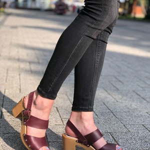 Puede incluir: Sandalias de tac&oacute;n de cuero burdeos con plataforma de madera y cierre de hebilla. Las sandalias tienen dos tiras sobre el pie y un dedo del pie abierto.
