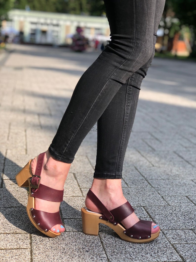 Puede incluir: Sandalias de tac&oacute;n de cuero burdeos con plataforma de madera y cierre de hebilla. Las sandalias tienen dos tiras sobre el pie y un dedo del pie abierto.