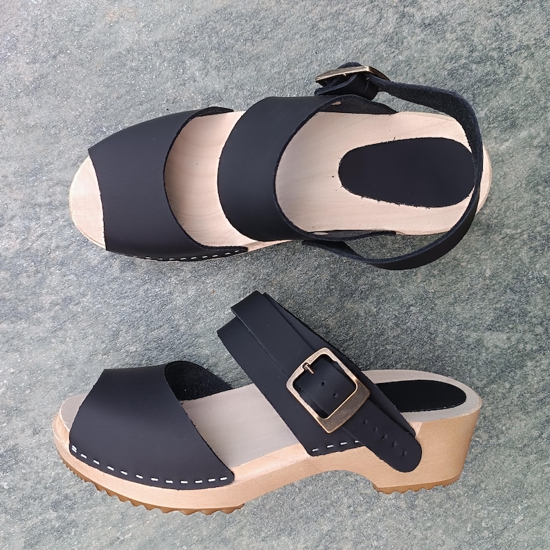 May include: A pair of black leather clogs with a wooden sole and a buckle strap. The clogs have a peep toe and a two-strap design.