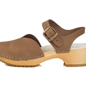 May include: A brown leather clog sandal with a wooden sole. The sandal features a closed toe, an ankle strap with a buckle, and a 2-inch heel. The upper is a light brown color, and the sole is made of light-colored wood.