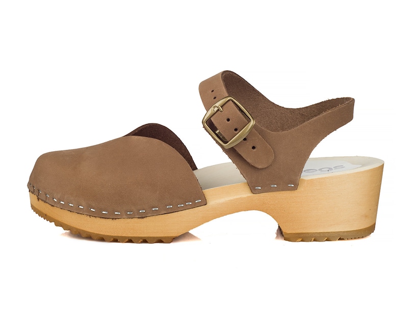 May include: A brown leather clog sandal with a wooden sole. The sandal features a closed toe, an ankle strap with a buckle, and a 2-inch heel. The upper is a light brown color, and the sole is made of light-colored wood.