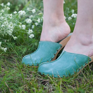May include: Teal suede clogs with wooden soles and brass stud detailing. The clogs are set on grass, with white flowers in the background. The clogs have a heel height of approximately 5 cm.