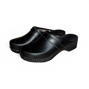 Pode incluir: Um par de tamancos de couro preto com cabedal liso, uma tira larga no peito do pé e uma sola grossa e escura. Os sapatos são construídos com costuras brancas visíveis ao longo da base, onde o cabedal encontra a sola, proporcionando um design limpo e funcional.