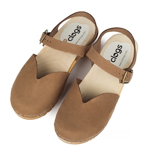May include: A pair of brown leather clogs with a wooden sole and a strap that buckles. The clogs have a closed toe design and a small heel. The brand name "clogs" is printed on the insole.