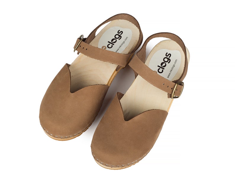 May include: A pair of brown leather clogs with a wooden sole and a strap that buckles. The clogs have a closed toe design and a small heel. The brand name "clogs" is printed on the insole.