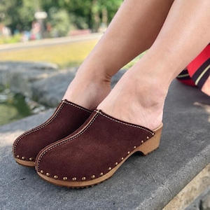 May include: A pair of dark brown suede clogs featuring a rounded toe, a light-coloured wooden sole, and a row of small metallic studs securing the upper material to the base. The footwear has a slip-on design with visible stitching along the centre seam.