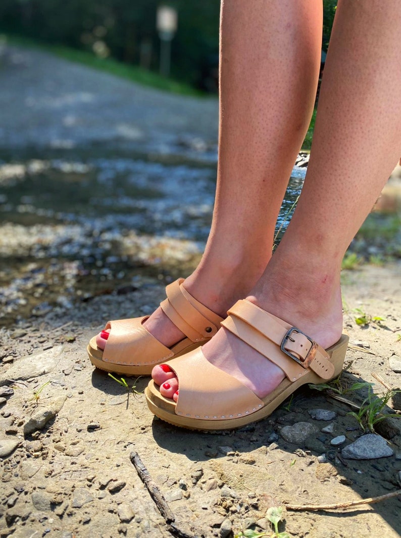 May include: A pair of tan leather sandals with a buckle closure. The sandals have a wooden platform sole and a peep toe design. The sandals are worn by a person standing on a dirt path.
