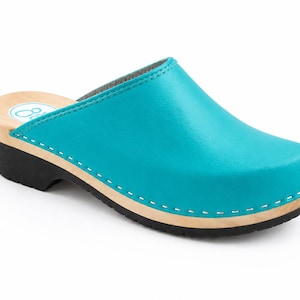 May include: A turquoise leather clog featuring a light-coloured wooden midsole and a black rubber outsole. The upper is attached to the wooden base with white stitching. A circular logo with a white footprint icon is visible on the inner heel.