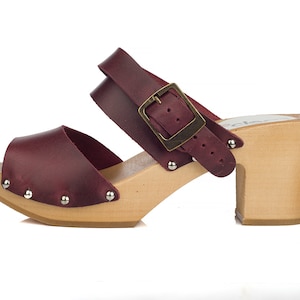 Puede incluir: Sandalias de madera con tac&oacute;n de cuero burdeos con una correa de hebilla y detalles de tachuelas. El zueco tiene una plataforma y un tac&oacute;n de madera.