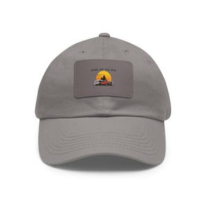 Peut inclure: Casquette de baseball grise avec une visière incurvée. Le chapeau présente un écusson rectangulaire avec le texte "KING OF MY RIG" au-dessus d'un dessin d'une caravane et d'une voiture rouge sur fond de coucher de soleil.