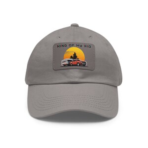 May include: A grey baseball cap featuring a rectangular patch on the front. The patch displays a graphic of an orange pickup truck towing a white camper trailer in front of a large yellow sun and black pine trees, with the text KING OF MY RIG.