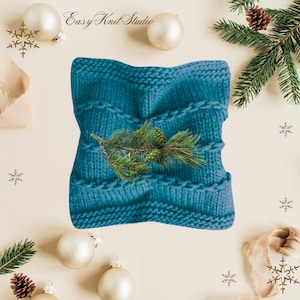 May include: A teal blue knitted pillow with a decorative stitch pattern. The pillow is surrounded by white ornaments and a pine branch with pine cones.