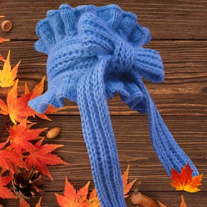 May include: A light blue knitted hat with a frilled edge and long ties. The hat is made from a soft, textured yarn, suitable for cold weather. The hat is displayed against a wooden background with autumn leaves.