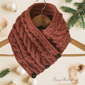 May include: A rust-coloured, knitted cowl with a cable knit design and three dark buttons. The cowl is displayed on a wooden hanger, with festive decorations in the background. The Easy Knit Studio logo is visible.
