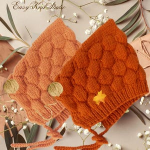 May include: Two knitted baby hats in orange and peach colors with a textured design. The hats have pom-poms and ties.