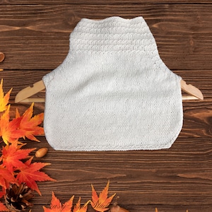 May include: A white knitted cowl neck warmer displayed on a wooden hanger. The neck warmer has a textured pattern at the top and a smooth knit body. The background features a wooden surface with autumn leaves and a pine cone.