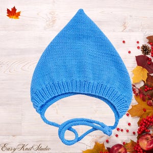 May include: A bright blue knitted bonnet with a pointed top and a ribbed brim. The hat features two long knitted ties extending from the base, designed to be fastened under the chin. The texture shows a consistent stockinette stitch pattern throughout.