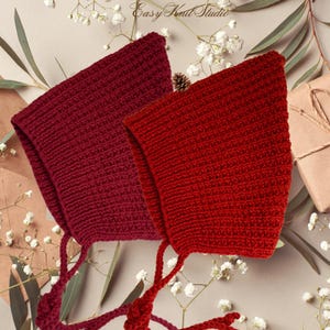 May include: Two pointed bonnets in deep burgundy and bright red, both featuring a ribbed knit texture and matching chin ties. The bonnets have a distinct triangular shape and are crafted with a consistent knit pattern throughout the fabric.