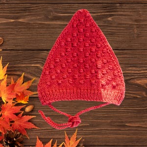 May include: A vibrant red knitted hat with a textured pattern and ear flaps. The hat is tied with a matching red string. The hat is likely for a baby or child. The hat is made of a soft, warm material.