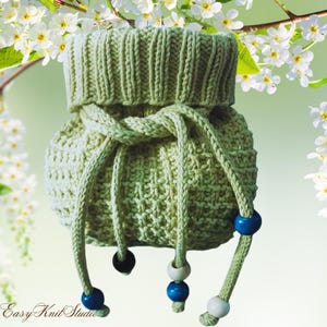 Könnte beinhalten: Ein hellgrüner Strickbeutel mit Kordelzug und strukturiertem Muster. Die Tasche hat einen gerippten oberen Teil und einen geflochtenen Kordelzug mit blauen, weißen und schwarzen Perlen. Die Tasche wird vor einem weichen grünen Hintergrund mit weißen Blumen präsentiert.