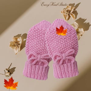 May include: A pair of pink knitted mittens with a bow detail. Each mitten has a small orange and yellow leaf on the top.