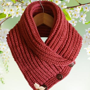 May include: A burgundy-colored, knitted cowl scarf with a ribbed texture. The scarf features two brown buttons and a decorative butterfly button. The words "Easy Knit Studio" are visible in the upper left corner.