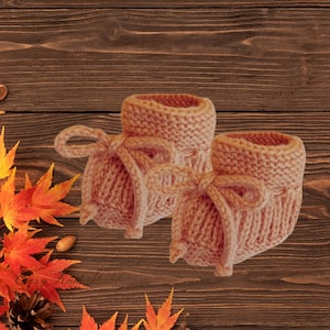 Pode incluir: Um par de botinhas de bebé de malha em tom pêssego suave. Cada botinha tem uma bainha enrolada e é amarrada com um laço de fio a condizer. As botinhas são exibidas numa superfície de madeira com folhas de outono.