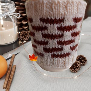 May include: A cream-colored knitted mug cozy featuring a repeating geometric pattern in dark rust brown. The cozy is crafted with a ribbed top edge and a textured knit stitch, designed to fit snugly around a cylindrical mug for insulation.