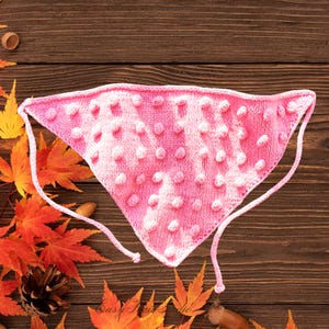 May include: A pink knitted bandana with a textured, dotted pattern. The bandana is triangular, with ties at the corners. The background features autumn leaves and a wooden surface.