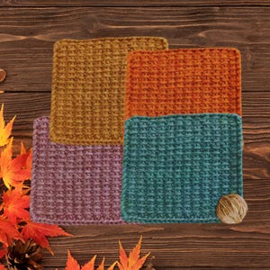 May include: Three square crochet washcloths featuring a textured waffle stitch pattern. The cloths are colored in mustard yellow, muted purple, and a two-toned combination of orange and teal, with a small ball of yarn attached to the teal section.